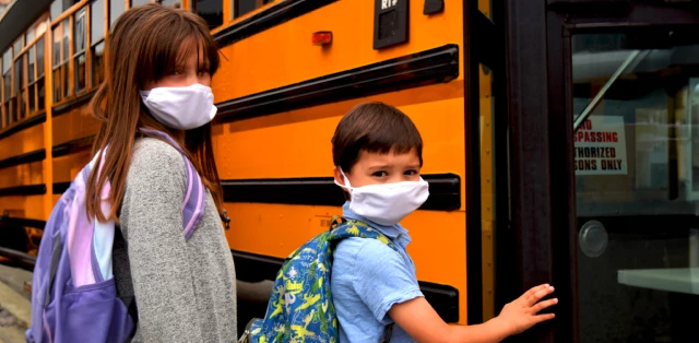 Two children in face masks stand outside a school bus, entering it.