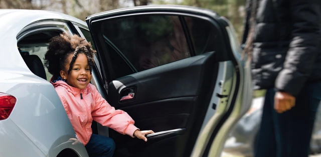 A child exciting a car smiling while an adult waits beside the car.
