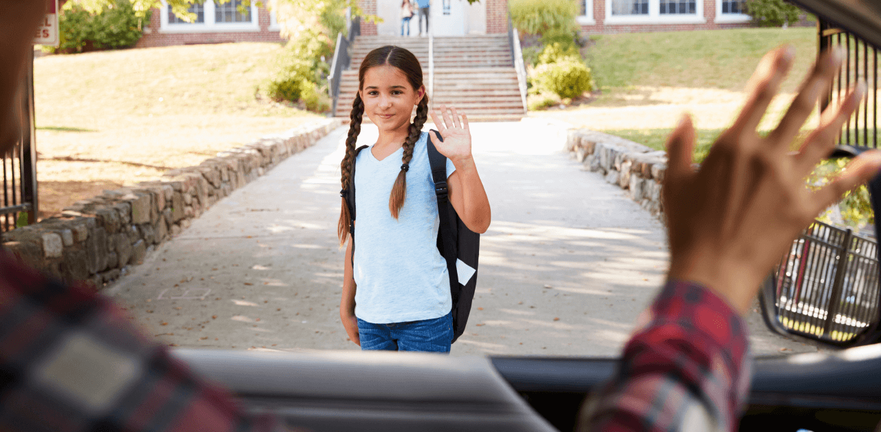 A young girl standing in front of a school, waves to a person from the window of a car.