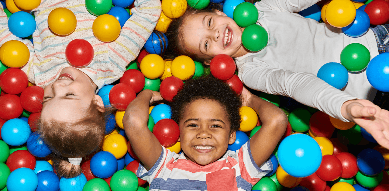Three cheerful children are beaming with smiles as they play together in a vibrant ball pit filled with assorted colored balls.