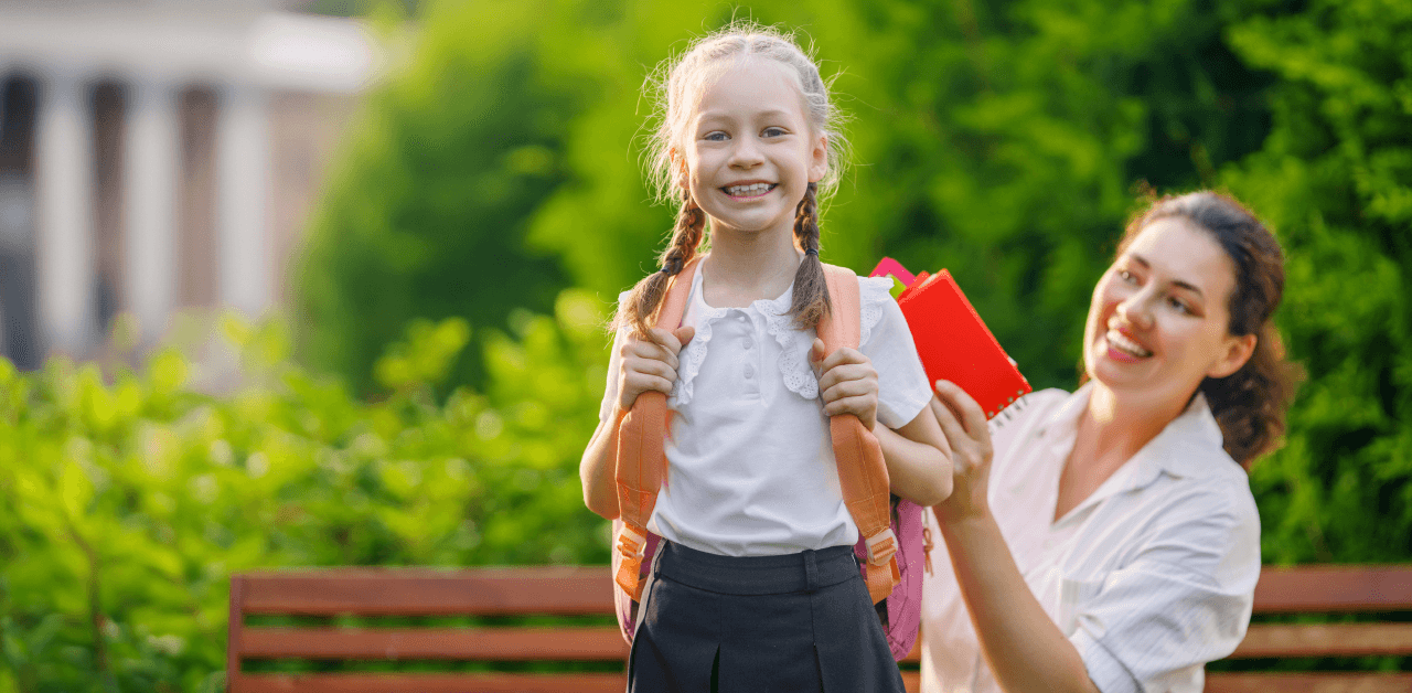 A woman stands behind a girl, putting school items in the child's backpack.