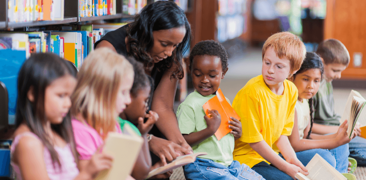 A woman reads a book to a group of attentive children in a library setting, surrounded by shelves of books.