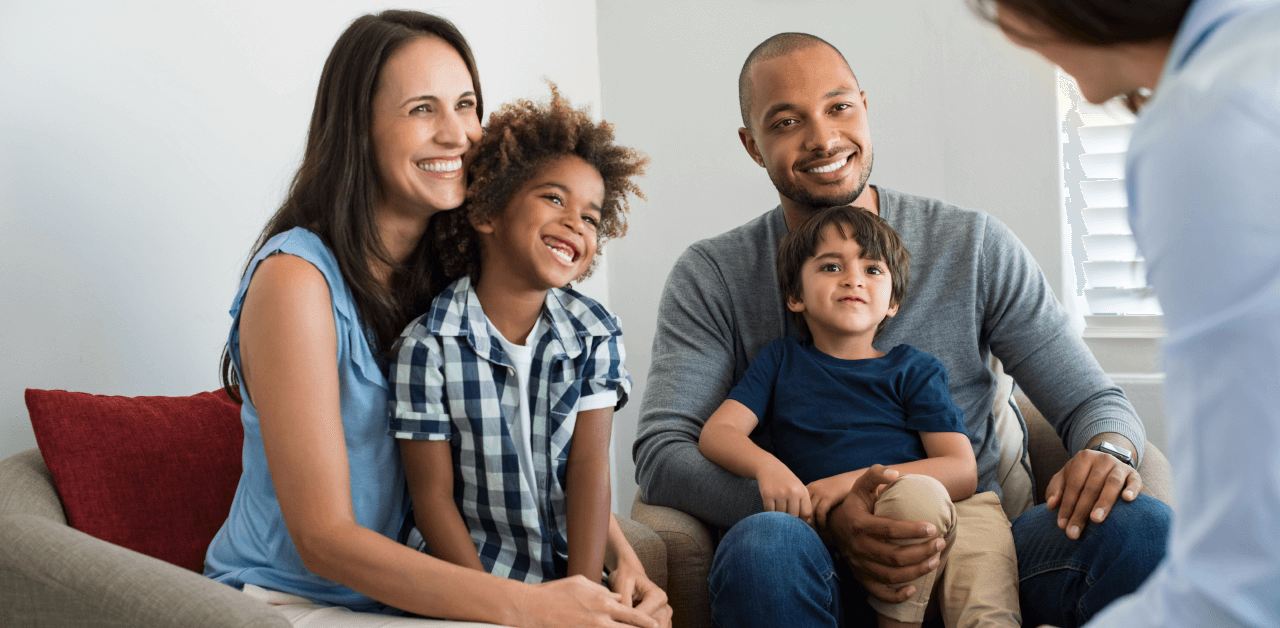 A family of four sits together on a couch, smiling at a person seated ahead of them.