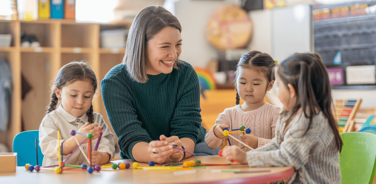 A smiling woman with three children at a round table in a colorful classroom setting, while the children plays with their learning tools.