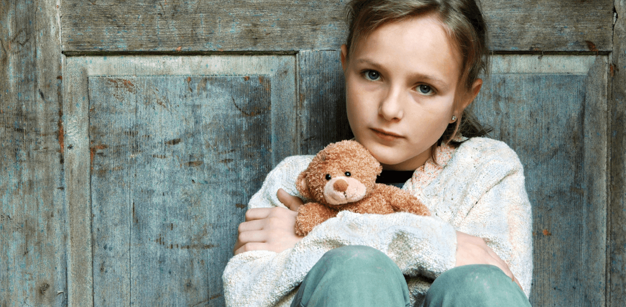 A young girl with a sad expression sits against a wooden door, holding a teddy bear in her arms.