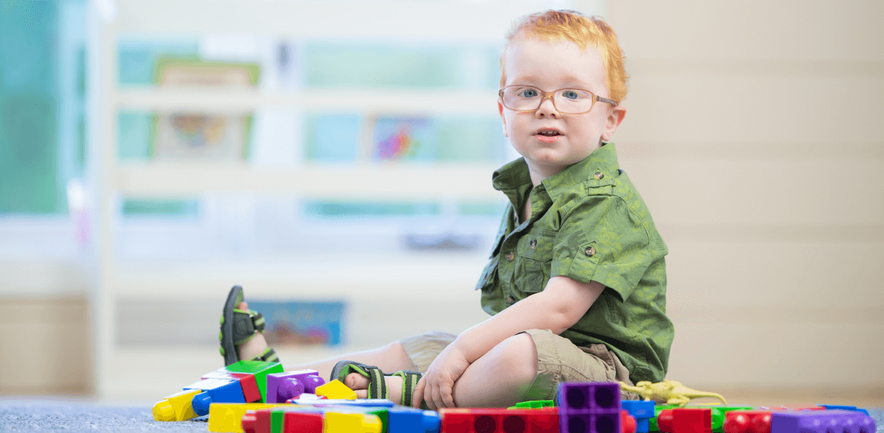 A young boy wearing glasses sits on the floor, surrounded by colorful building blocks.