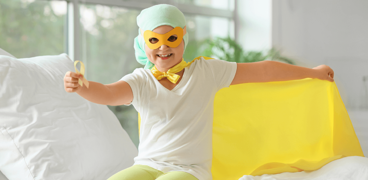 A young child in a bright yellow cape and mask sits on a bed, radiating joy and imagination in a playful setting.