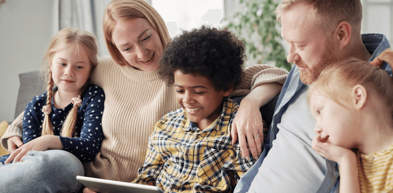 A family of four sits together on a couch, engaging with a tablet, sharing smiles and enjoying quality time.