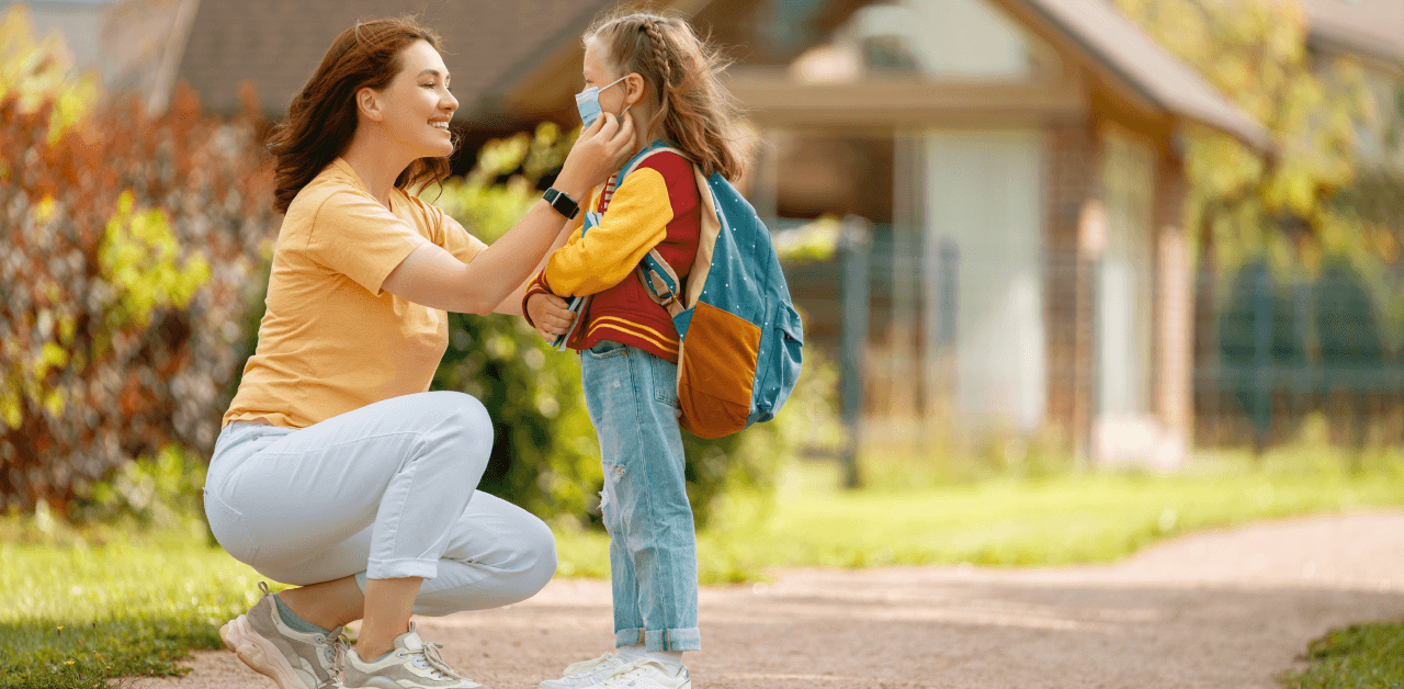 A woman kneels on the sidewalk, adjusting a young girl's mask.
