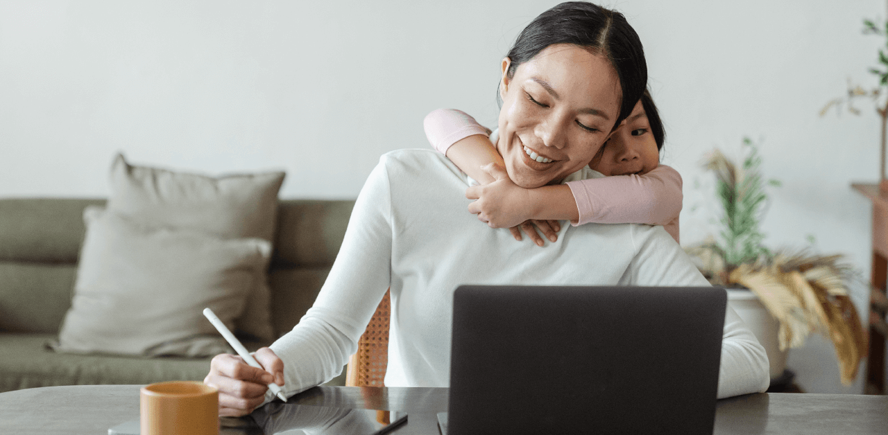 A smiling mother works in front of a laptop while her child envelopes her in a big hug.
