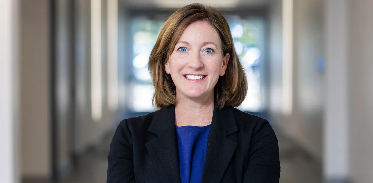 Bridgette Brinkmann, Chief People Officer, smiling confidently with arms crossed, wearing a professional black blazer and blue top in a bright office hallway