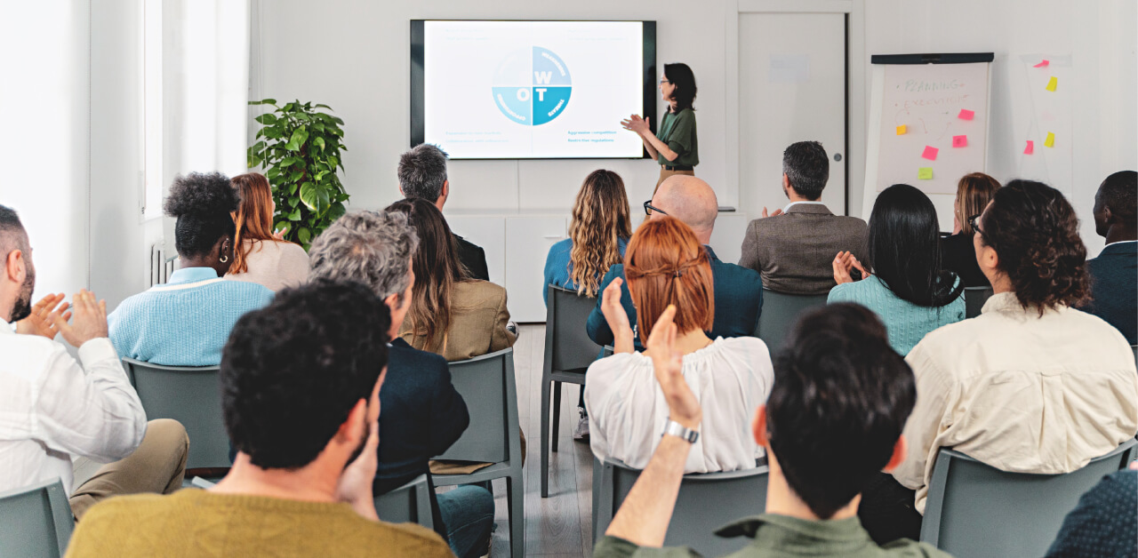 A group of people watching a presenter in a conference room, with a presentation on a screen in the background