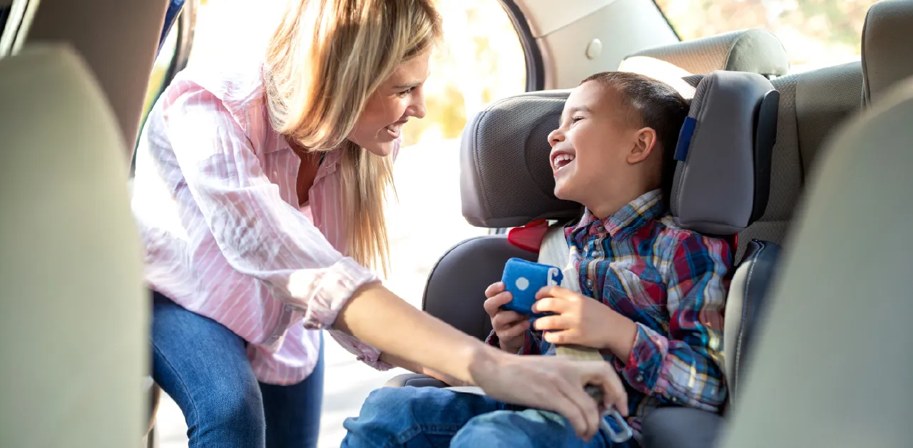 A woman smiles while carefully buckling a young child into his car seat
