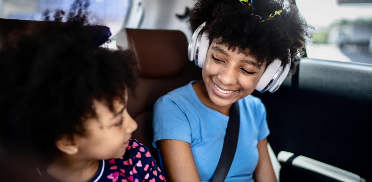 Two young girls sit in the back seat of a car, smiling, with one wearing headphones and seatbelt.