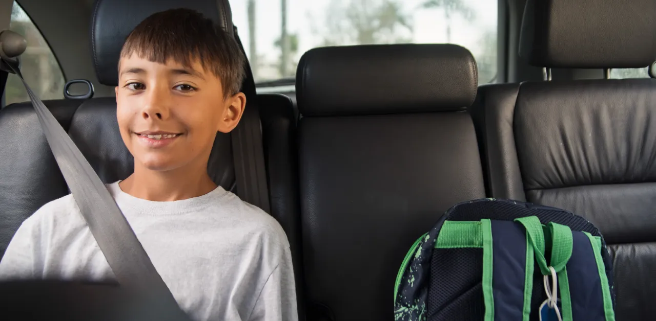 A schoolboy smiles as he sits buckled in the backseat of a car next to a blue and green backpack.
