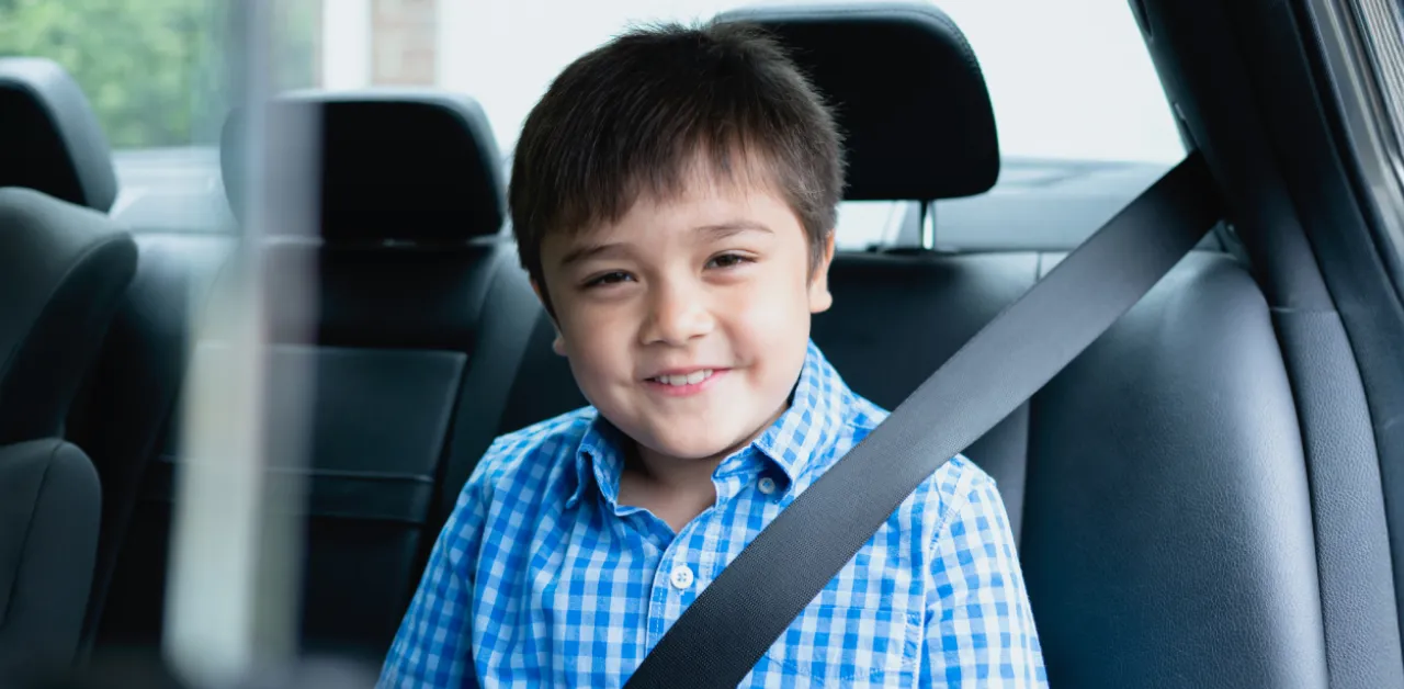 A schoolboy in a blue checkered shirt smiles as he sits securely in a car's back seat, properly fastened with a seatbelt
