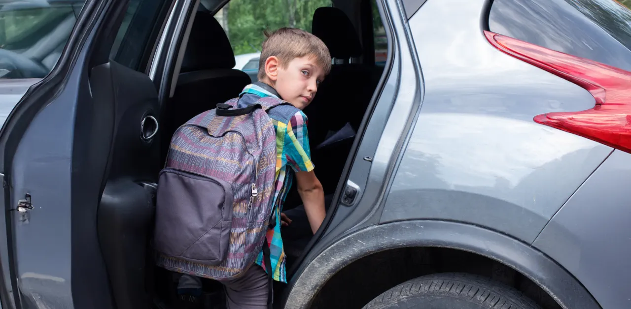 Young schoolboy in plaid shirt and backpack climbs into the back seat of a gray car.