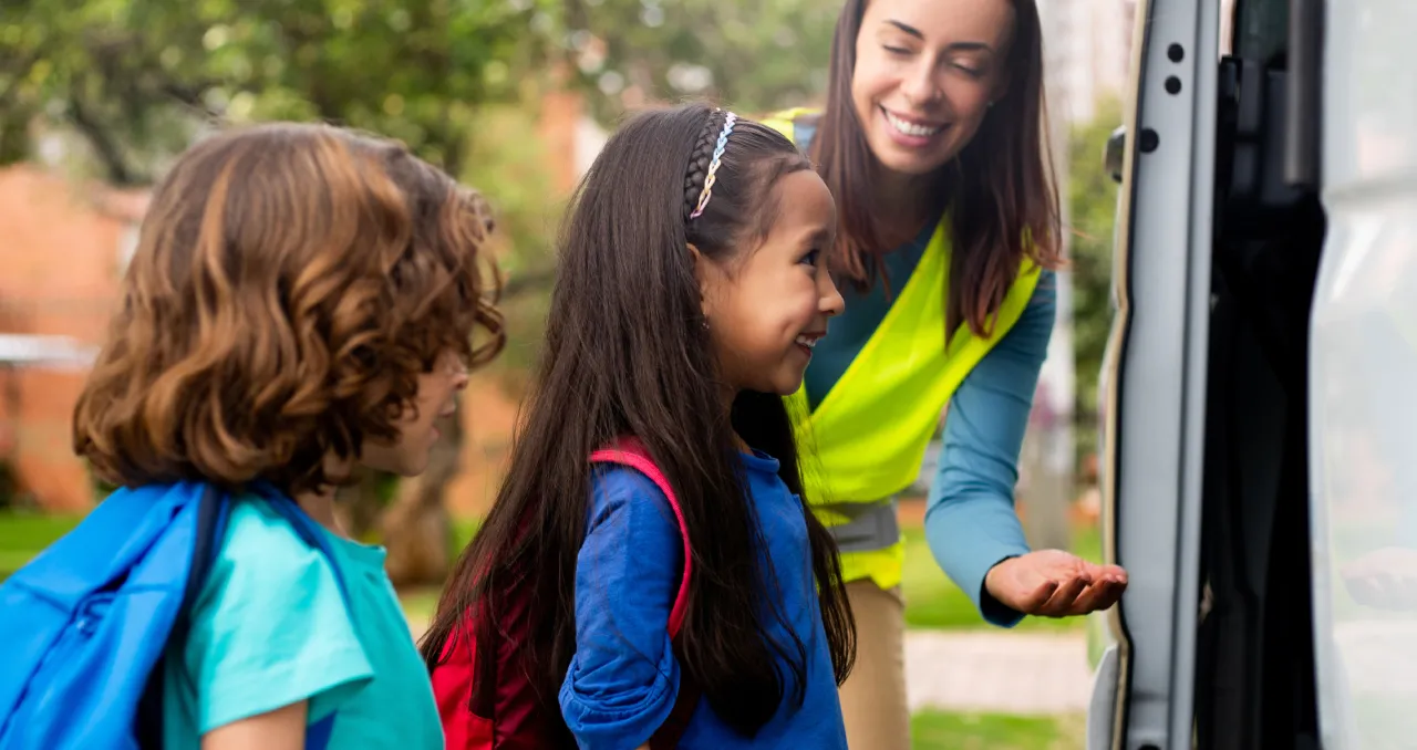 Two schoolchildren with backpacks prepare to board a vehicle. An adult in a high-visibility vest guides them.