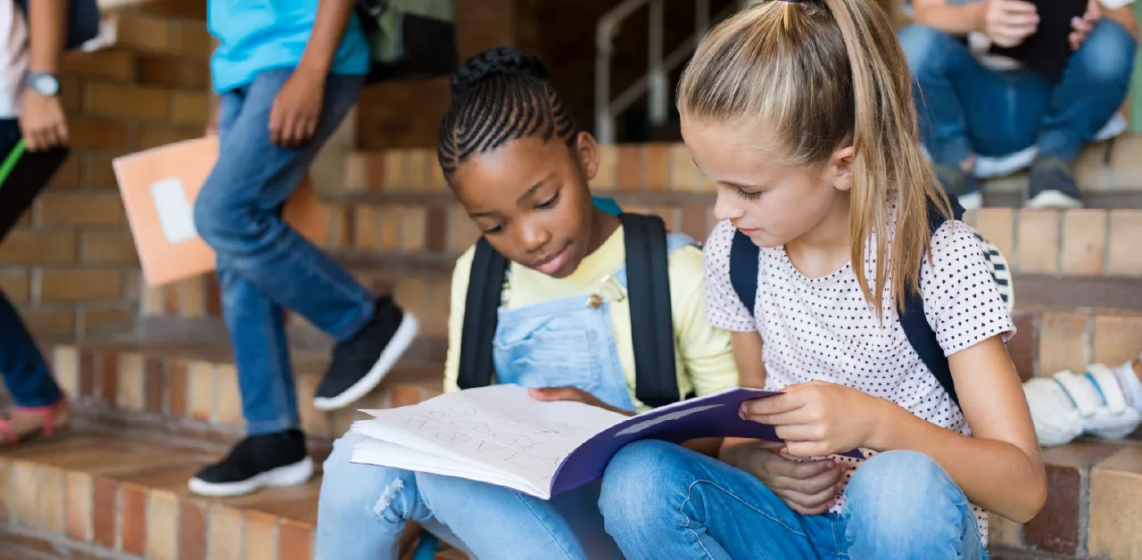 Two elementary school girls reading a workbook together on school steps