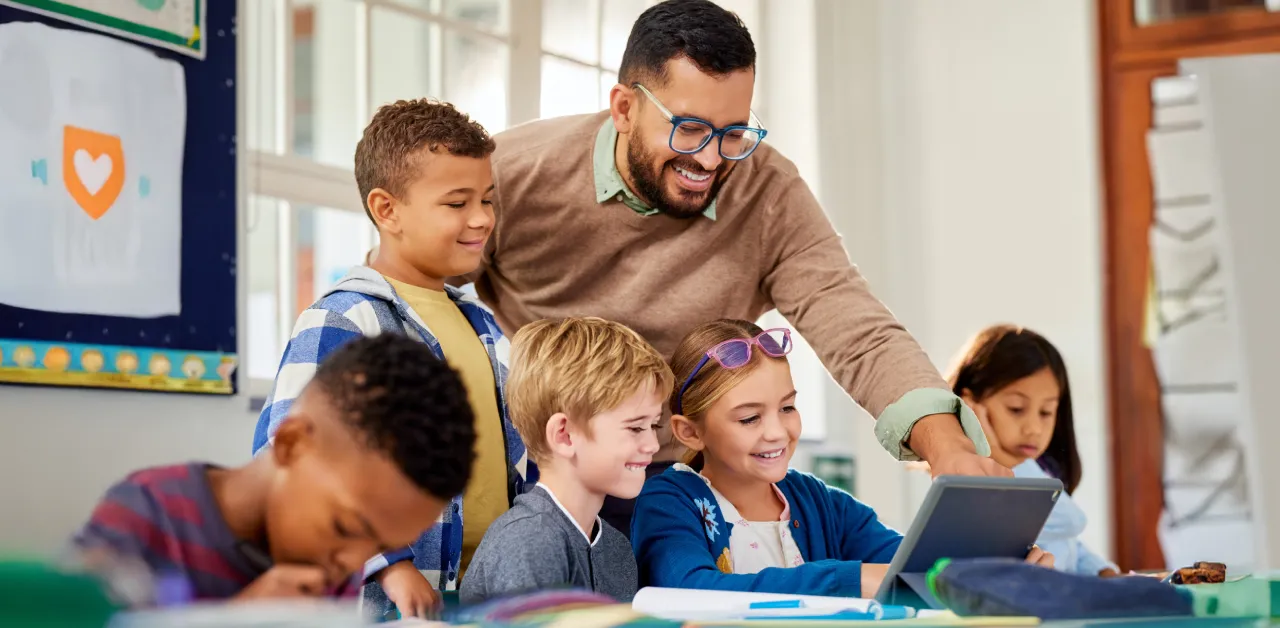 acher guides young schoolchildren around a tablet in a bright classroom.
