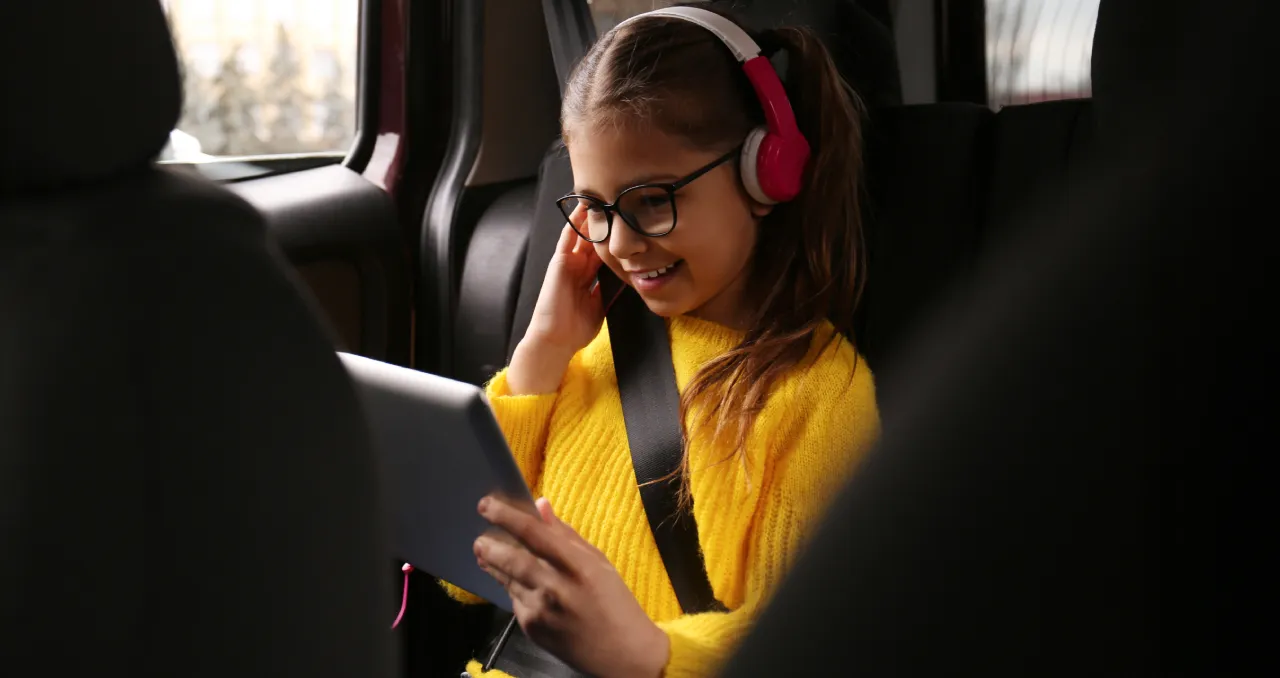A young girl wearing glasses and pink headphones smiles while watching a tablet in the backseat of a car, safely buckled in with a seatbelt.