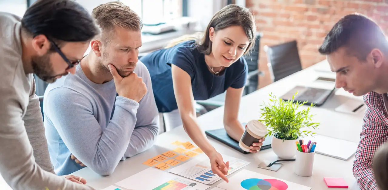 A diverse team collaborates around a table with charts, graphs, and sticky notes. Office supplies, a plant, and coffee set a casual work vibe.