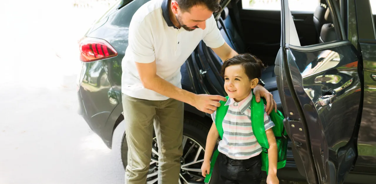 An adult helps a schoolboy adjust their green backpack by an open car door.