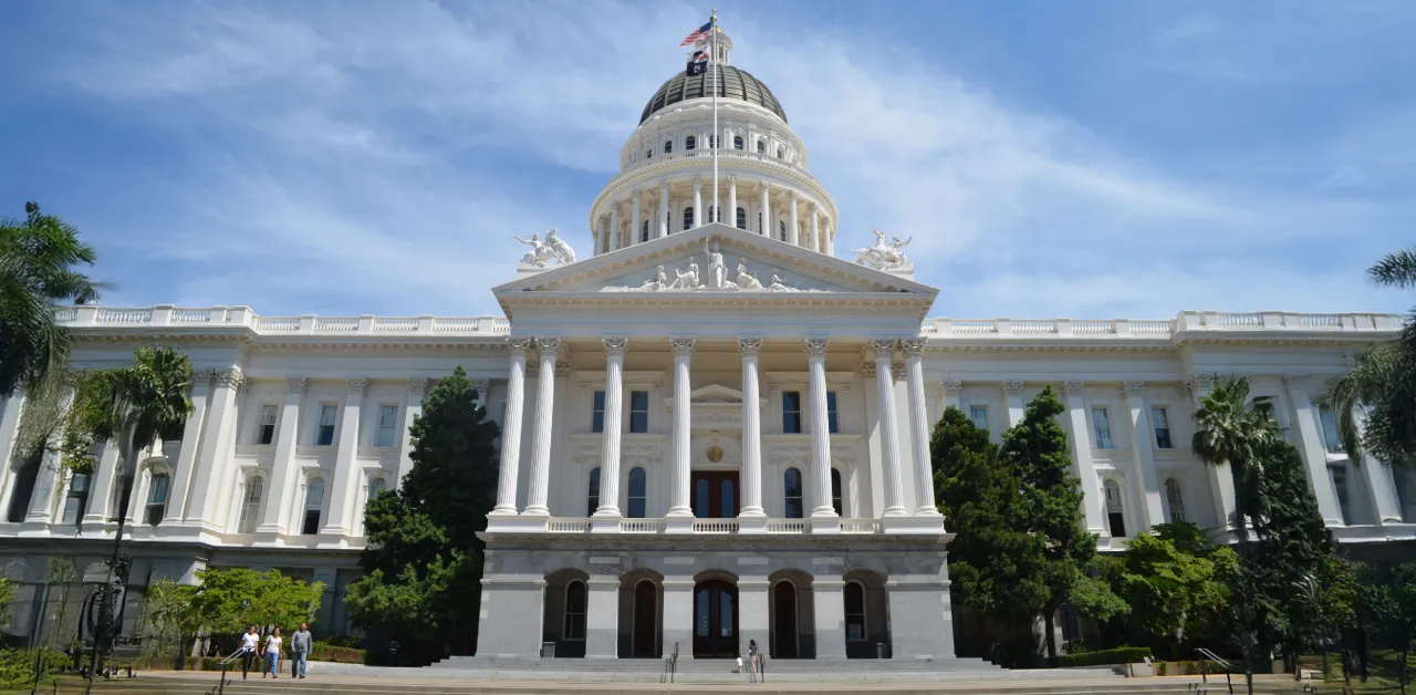 The front exterior of the California State Capitol building in Sacramento, featuring columns, a dome, and surrounding greenery under a blue sky.