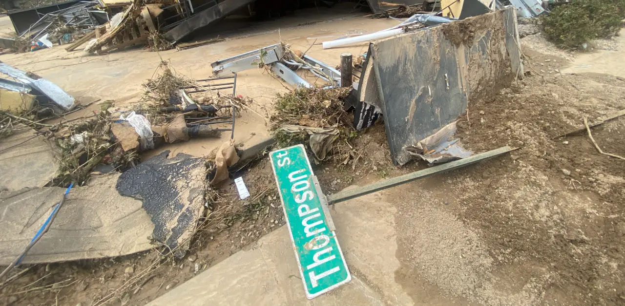 A fallen 'Thompson St.' sign lies amidst debris and mud after a severe disaster.