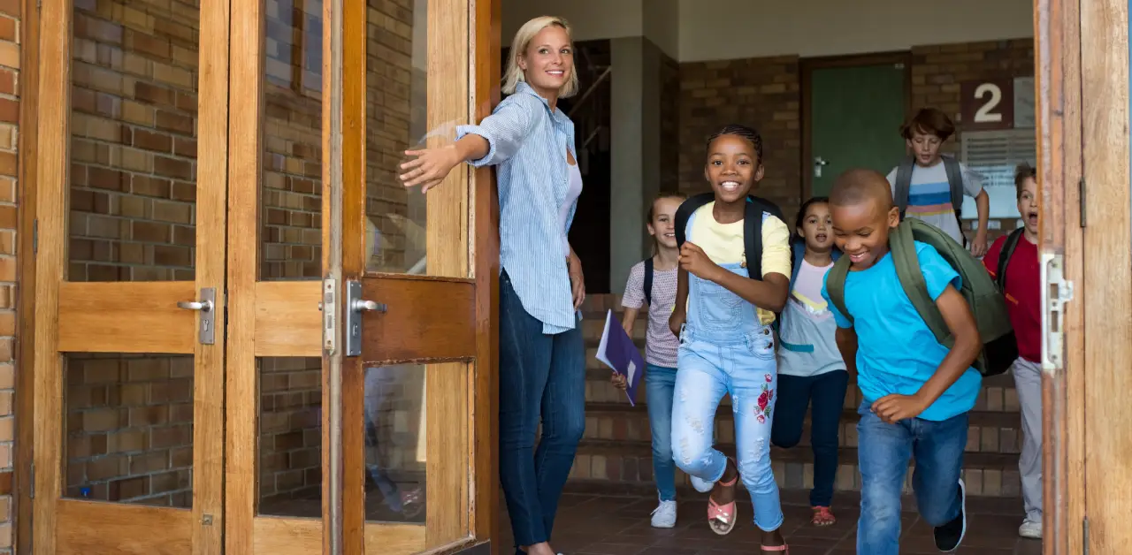 Children exit a school building excitedly with backpacks and notebooks as a staff member holds the door open.