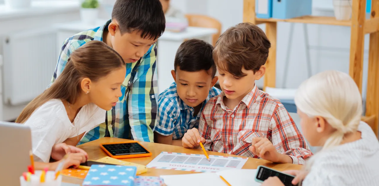 Five elementary school students gathered around a table working together on a school project with pencils, tablets, and a printed chart.