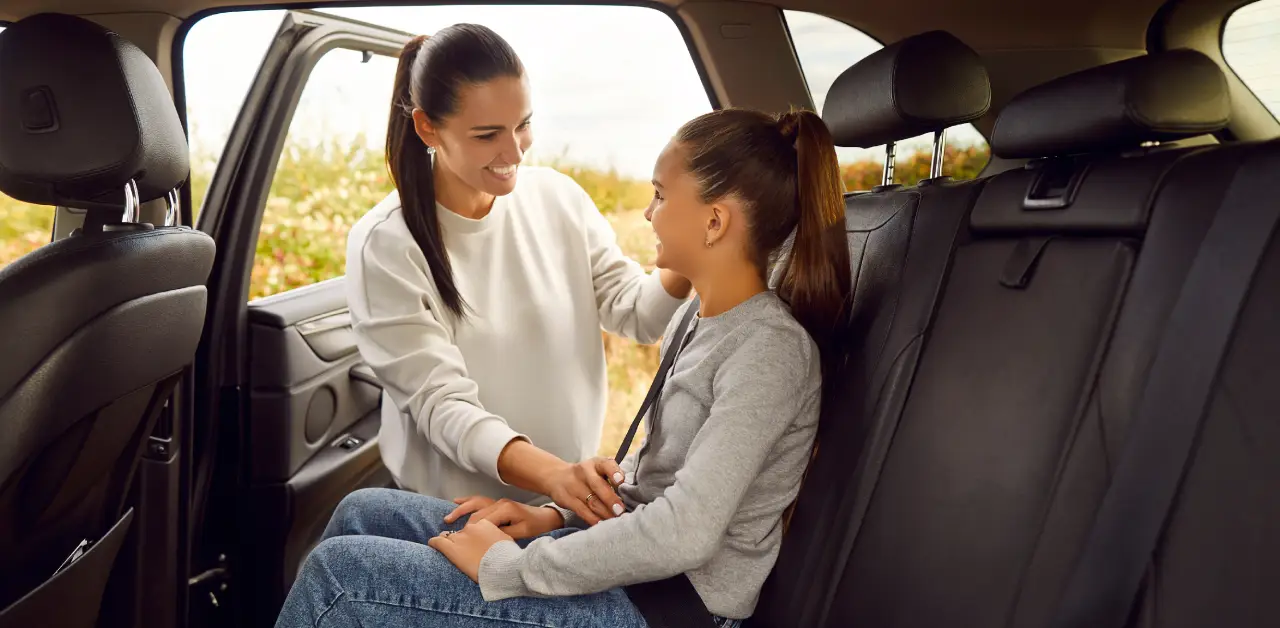 A woman helps a young girl buckle her seatbelt in the backseat of a car. The open door reveals a scenic outdoor.
