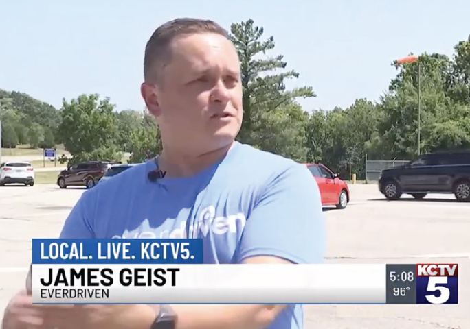 KCTV5 interviews EverDriven’s James Geist, wearing a blue EverDriven shirt, outdoors in a parking lot with cars and trees in the background on a windy day.