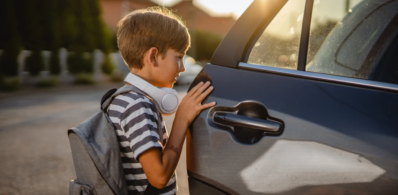 A young schoolboy wearing a backpack and headphones around his neck opens the car door while standing next to the vehicle in warm sunlight.