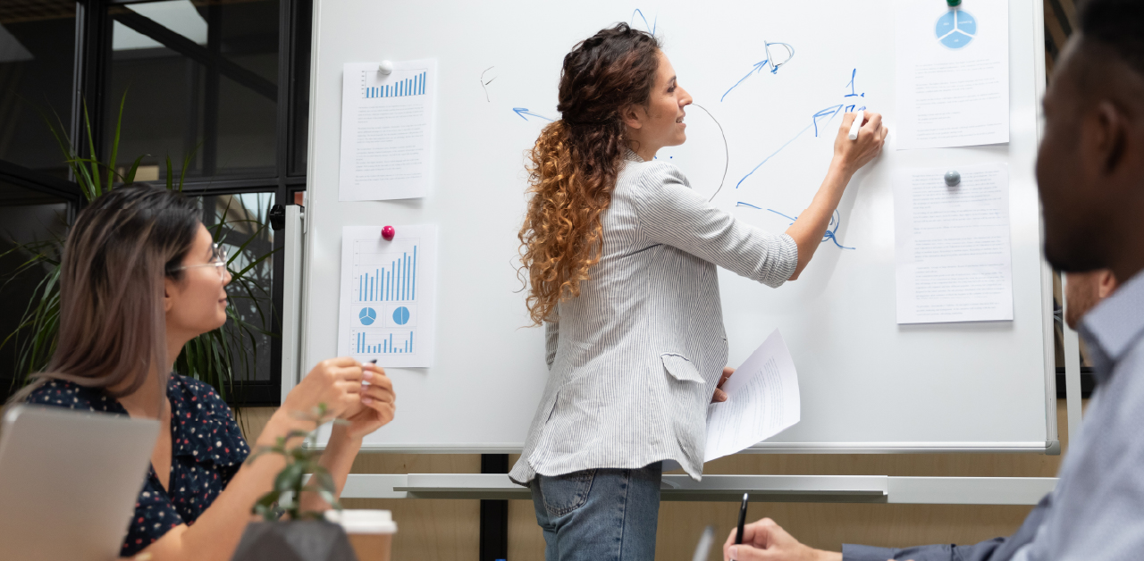 A woman presents ideas on a whiteboard to engaged colleagues who are seated nearby, taking notes in an office brainstorming session
