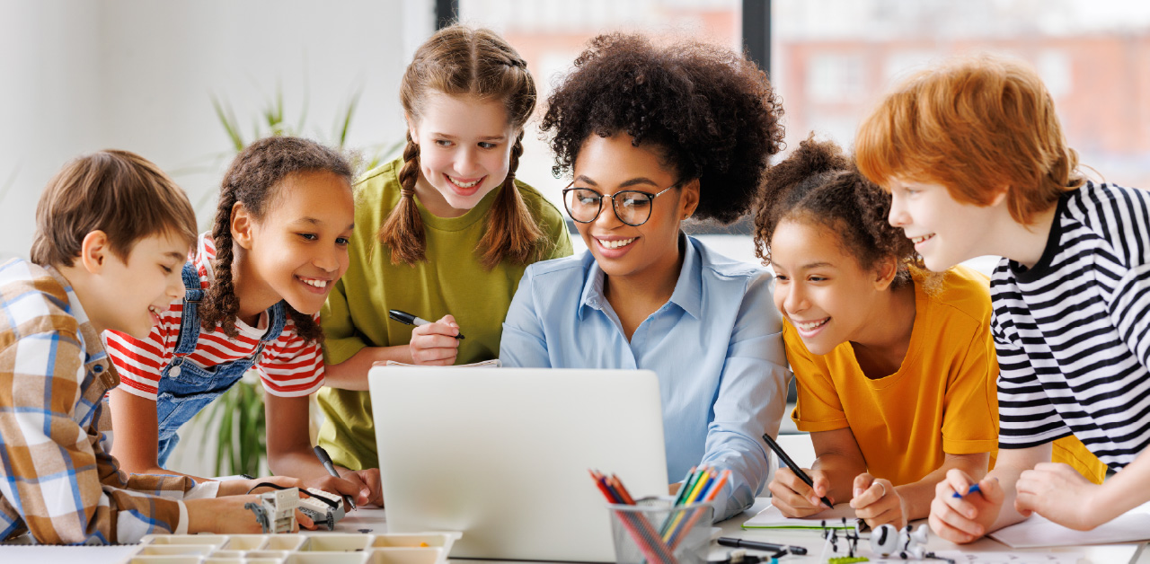 students gather around a teacher using a laptop during a classroom activity