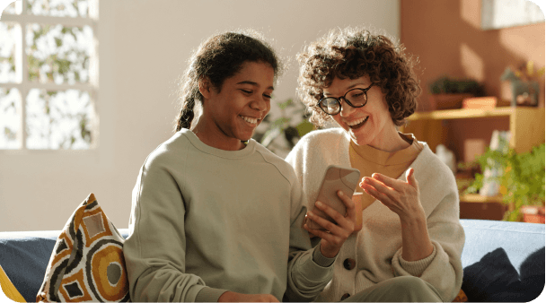 A smiling young person and an adult with curly hair and glasses sit on a couch, sharing a joyful moment over a smartphone.