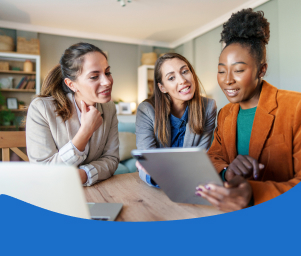 Three women in a cozy office setting are smiling and discussing something on a tablet. They exude enthusiasm and collaboration. Warm lighting enhances a welcoming mood.