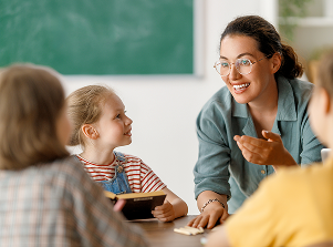 A smiling teacher engages with three young children in a classroom, discussing happily. The teacher's hand is raised as if making a point.