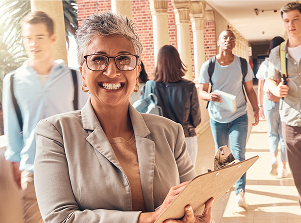 Smiling woman with short gray hair and glasses holding a clipboard stands in a school hallway. Students walk by, creating a lively atmosphere.