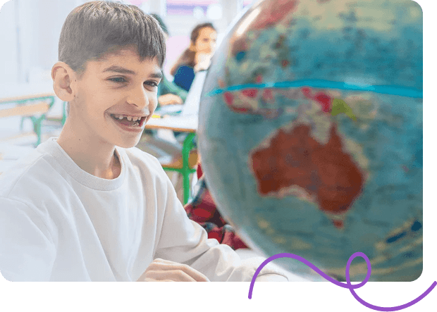 A boy in a white shirt smiles while looking at a large globe in a classroom.