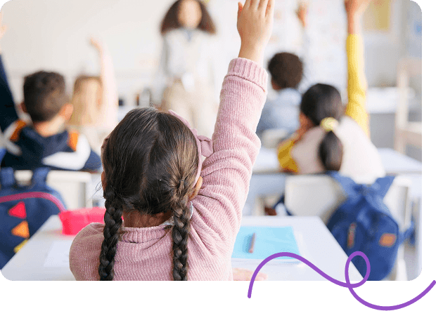 In a classroom, a young girl with braided hair raises her hand enthusiastically.