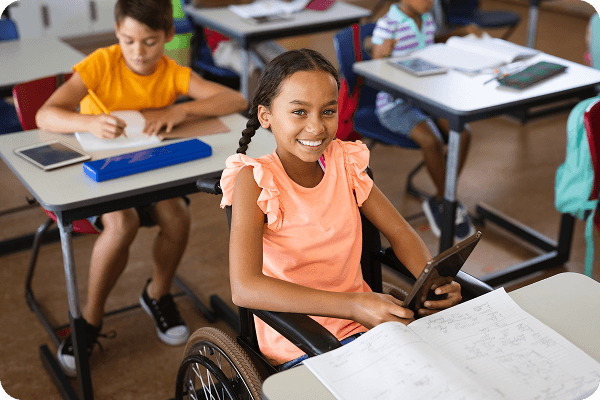 A smiling girl in a wheelchair holds a tablet in a classroom. She's wearing an orange top, surrounded by classmates at desks.