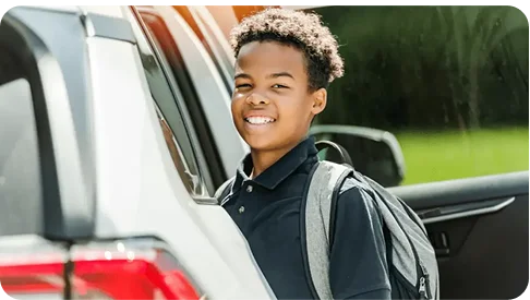 A smiling child with a backpack stands beside a white car with an open door.