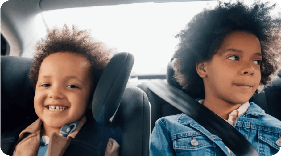 Two children with curly hair sit in the back seat of a car. The younger child smiles joyfully, while the older child looks forward with curiosity. Both wear seatbelts.