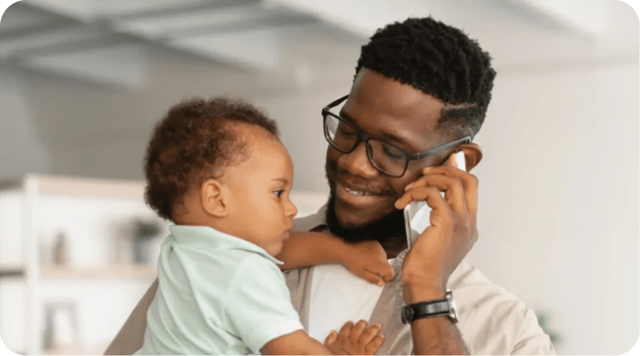 A smiling man wearing glasses talks on the phone while holding a baby. The bright, modern room in the background suggests a warm and happy environment.