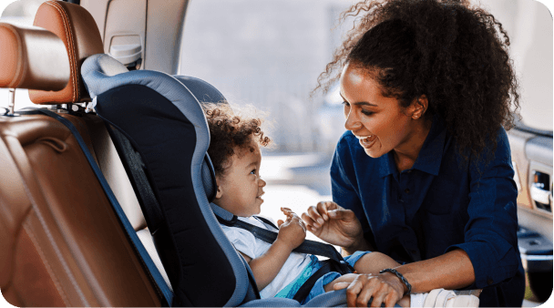 A smiling woman kneels beside a car seat, engaging with a happy toddler inside a vehicle.